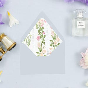 May include: A light blue envelope with a floral patterned interior. The interior design features pink and peach flowers with green leaves on a white background. The envelope is surrounded by flowers, a perfume bottle, and a decorative swan.