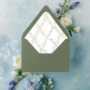May include: An olive green envelope with a floral patterned interior, featuring a delicate arrangement of pink, blue, and yellow flowers. The envelope is set against a light blue, textured background with white and blue flowers.