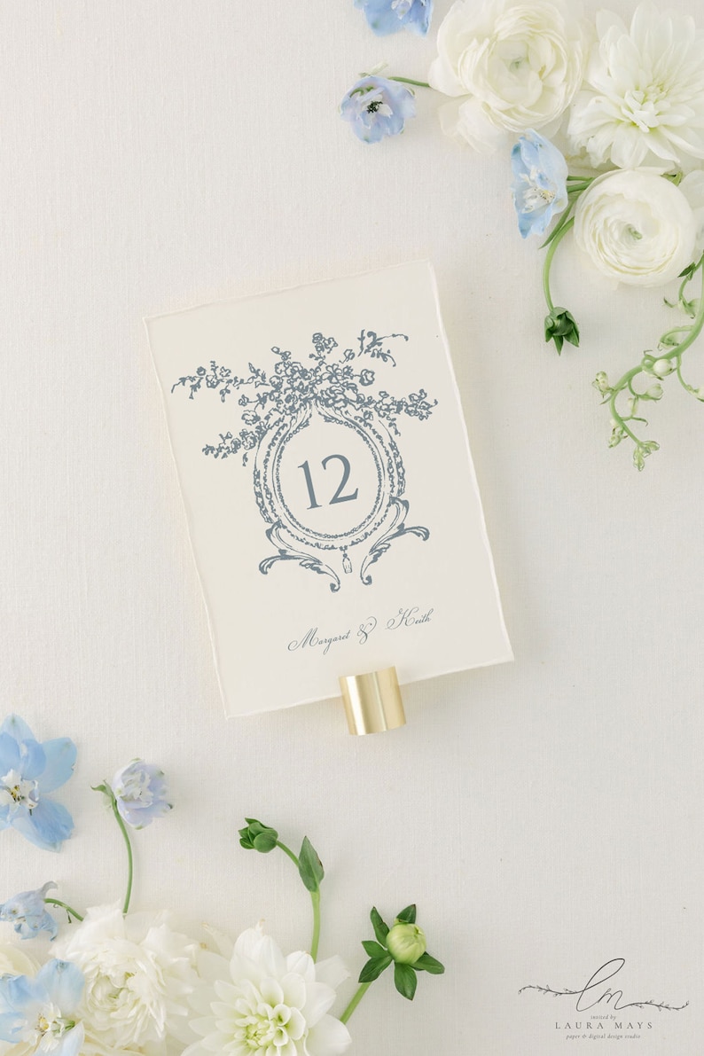 May include: A white card with a blue floral design and the number 12 in a circle. The card also has the text "Margaret & Keith" written in cursive. The card is attached to a gold metal stand.