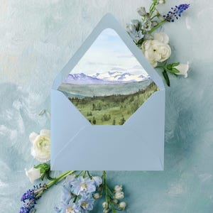 May include: A light blue envelope with a watercolor landscape of mountains and trees. The envelope is open, revealing the artwork. The image is surrounded by white and blue flowers. Text at the top reads "shown in azure blue".