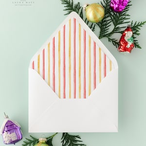May include: A white envelope with a striped interior design in red and gold. The envelope is surrounded by Christmas ornaments and evergreen branches, creating a festive holiday theme. The top left corner has the text "Laura Mays" in script.