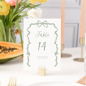 May include: A white table number card with a green ribbon bow design. The card reads "Table 14" and "Emily & Michael March 18, 2025".