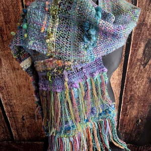 May include: A hand-woven scarf featuring a multi-colored palette of blue, purple, green, and orange yarns. The textile displays a complex, textured weave with integrated loops, knots, and a long, multi-strand fringe at the bottom edge.