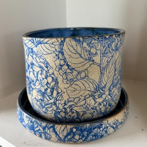 Puede incluir: Una maceta de cerámica con su platillo a juego. La maceta y el platillo están decorados con un estampado floral azul sobre un fondo crema. Los bordes son de color azul. La maceta está diseñada para plantas. Diámetro aproximado de 15 cm.