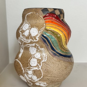 May include: A speckled tan ceramic vase featuring two white skull illustrations on the left side and a raised, glossy, multi-colored rainbow design on the right. The top rim of the vase displays a black and tan checkered pattern.