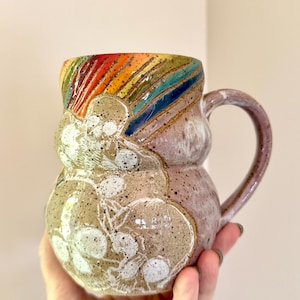 May include: A ceramic mug with a rounded, bulbous shape and a curved handle. The upper rim features a vibrant, multi-colored rainbow glaze. The body has a speckled beige finish with a white, etched botanical design of leaves and berries.