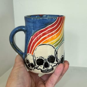May include: A ceramic mug with a blue glazed finish featuring a hand-painted rainbow arching over three white skulls with black outlines and details. The mug has a curved handle and a cylindrical shape, showcasing a contrast between the dark blue background and the vibrant colors of the rainbow design.