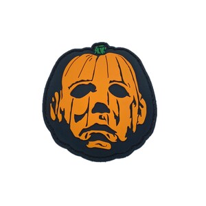 May include: A Halloween-themed patch featuring an orange pumpkin with a face resembling Michael Myers from the horror film series. The patch has a black border and a green stem, perfect for adding to clothing or accessories.