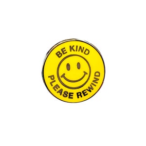 May include: Yellow enamel pin with a smiley face and the text "Be Kind Please Rewind".