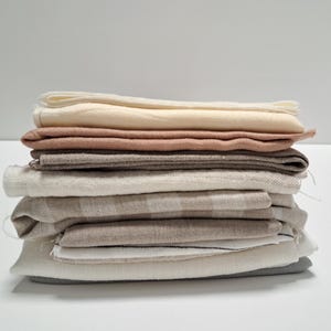 Op de afbeelding: Een stapel gevouwen stukken stof in diverse neutrale tinten, waaronder crème, beige, lichtbruin en zachtgrijs. De textielsoorten hebben verschillende texturen en wevingen, waarbij sommige subtiele streepjespatronen en rauwe randen tonen.
