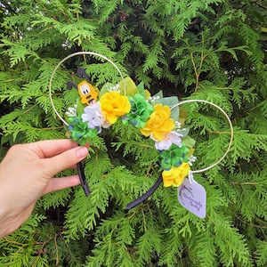 Puede incluir: Una diadema floral con orejas doradas de Mickey Mouse. La diadema presenta flores amarillas, verdes y blancas, y un personaje de Pluto. La diadema se sostiene frente a un fondo de hojas verdes. Se adjunta una etiqueta con texto.