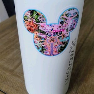 May include: White water bottle with a Mickey Mouse shaped sticker featuring a map of a theme park. The sticker has a blue outline and features a pink and purple color scheme. The text "#yesstraw" is printed on the bottle.