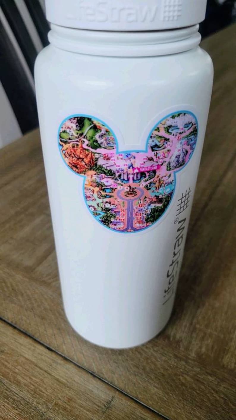 May include: White water bottle with a Mickey Mouse shaped sticker featuring a map of a theme park. The sticker has a blue outline and features a pink and purple color scheme. The text "#yesstraw" is printed on the bottle.