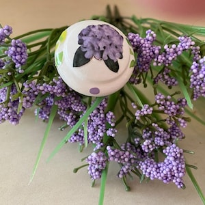 May include: White ceramic knob with a purple flower design and green leaves. The knob has small white polka dots around the edge.