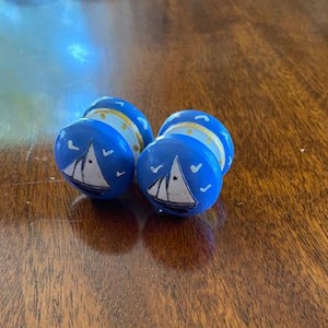 Peut inclure: Deux boutons de tiroir en bois bleu avec un motif de voilier blanc et des accents dorés. Les voiliers sont sur un fond bleu avec des oiseaux blancs.