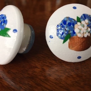 May include: Two white ceramic knobs with blue and white floral designs. The knobs have a blue and white polka dot pattern around the edges. The flowers are in a brown basket.
