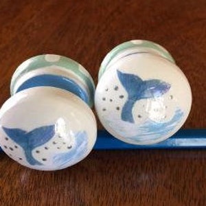 May include: Two white ceramic door knobs with a blue and green design. Each knob features a painted blue whale tail with white bubbles and a blue wave.