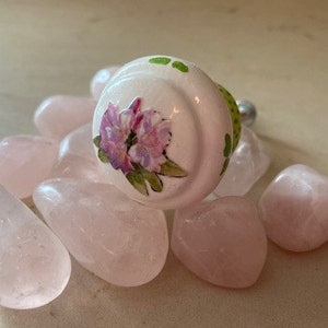 May include: A white ceramic knob with a floral design and green polka dots. The knob is surrounded by pink rose quartz stones.