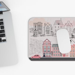 May include: A mouse pad with a whimsical illustration of a city street with houses, a car, and a bird. The background is a light pink with a watercolor effect.