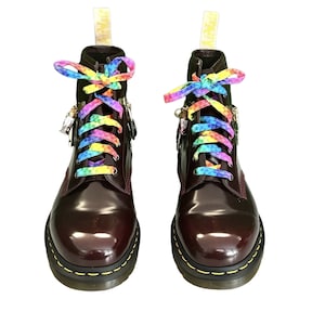 Puede incluir: Botas de cuero color burdeos con cordones arcoíris. Las botas tienen un acabado brillante, costuras amarillas y cremalleras laterales. Los cordones están atados con un lazo, sobre un fondo blanco.