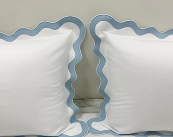 Pode incluir: Duas fronhas brancas com uma borda ondulada azul clara. As fronhas são feitas de um tecido liso e são exibidas lado a lado. A borda azul clara adiciona um toque decorativo à roupa de cama.