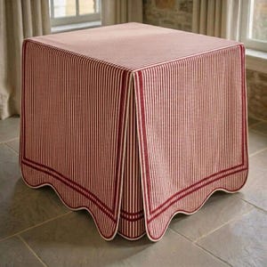 Puede incluir: Una mesa cuadrada cubierta por un mantel de tela con un patrón de finas rayas verticales rojas y blancas. El mantel tiene un borde festoneado rematado con una franja roja gruesa que recorre la parte inferior de los cuatro lados de la mesa.