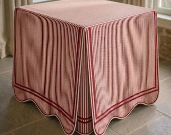 May include: A square table covered by a fabric cloth featuring a pattern of thin vertical red and white stripes. The cloth has a scalloped hemline finished with a thick red border that runs along the bottom edge of all four sides of the table.