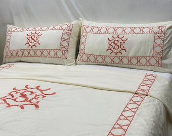 May include: Cream-colored bedding set with red embroidered details. The set includes two pillowcases and a duvet cover, all featuring a geometric border and a central monogram. The monogram is in a decorative font. The overall design is classic and elegant.