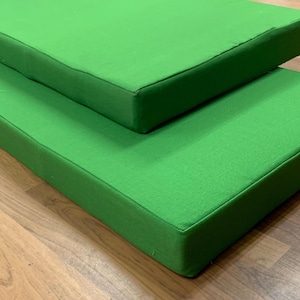 May include: Two bright green rectangular cushions are stacked on a wooden surface. The cushions have a simple, tailored design with a solid color and a clean, modern aesthetic. The cushions are likely for seating or decorative purposes.