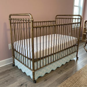 Könnte beinhalten: Ein Babybett im Vintage-Stil mit einem bronzefarbenen Metallrahmen. Das Bett hat vertikale Stäbe und eine weiße Matratze. Ein gemusterter blau-weißer Bettrock ist sichtbar. Das Bett steht auf Rollen.