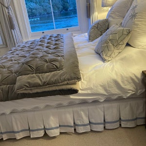 Può includere: Un letto bianco con un copriletto trapuntato grigio e cuscini con motivi bianchi e grigi. Il letto ha una gonna da letto bianca con un bordo blu. Il letto si trova in una camera da letto con una finestra che si affaccia su uno specchio d'acqua.