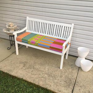 May include: White wooden bench with a colorful striped cushion. The bench has a slatted back and armrests. Two white planters are stacked nearby. A small decorative fountain sits to the left.