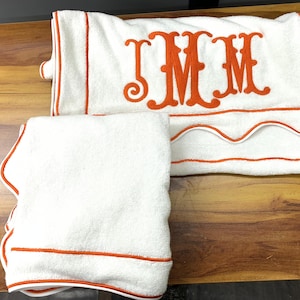 May include: White bath towels with orange trim and embroidered initials. One towel is folded, and the other is folded with a decorative edge. The towels are on a wooden surface.