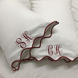Op de afbeelding: Twee witte kussenslopen met bordeaux geschulpte randen en geborduurde initialen. De kussenslopen liggen op een wit laken. Eén kussensloop heeft de initialen "SK" en de andere "CK".