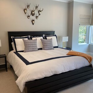 Può includere: Una struttura letto nera con un piumino bianco e un bordo blu scuro. Il letto ha un cuscino con motivo bianco e blu e un cuscino bianco con un bordo blu scuro. Ci sono diverse teste di cervo decorative sul muro dietro il letto.