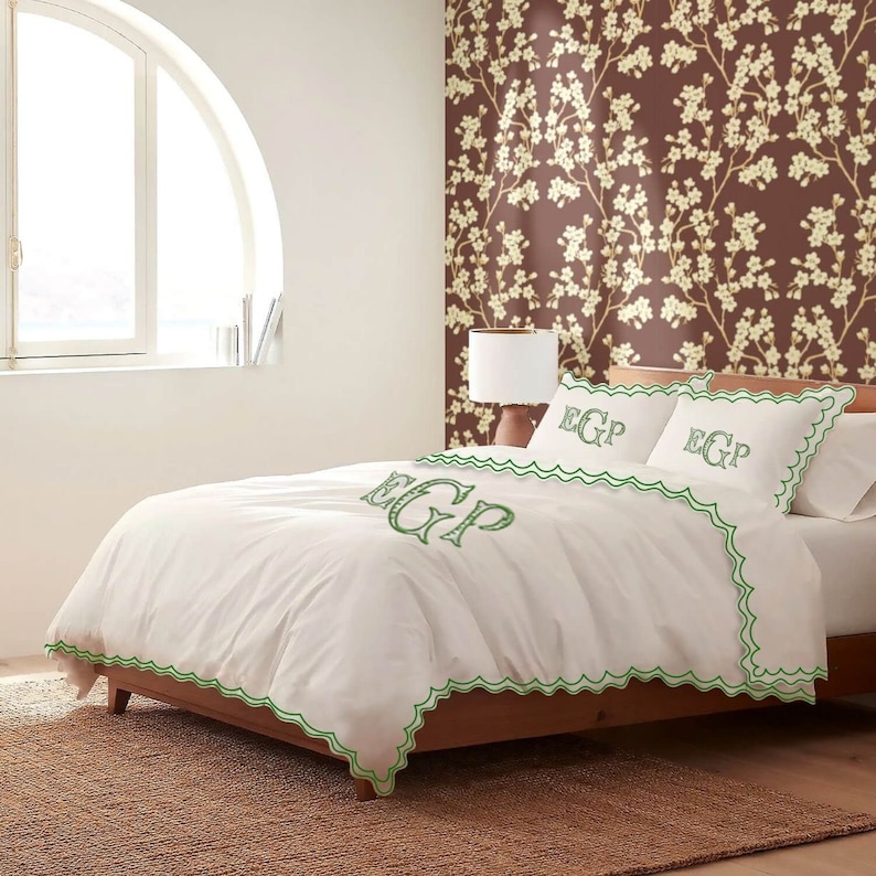 May include: White bedding with green scalloped trim and embroidered initials "EGP" on the duvet cover and pillowcases. The bedding is on a bed with a brown wooden frame and a brown floral patterned wallpaper.