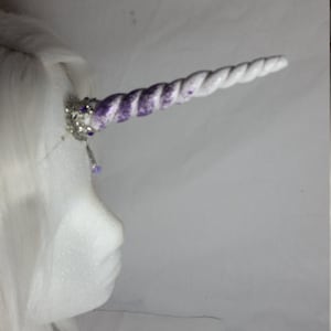 May include: A white unicorn horn with a purple and white swirl design. The horn is attached to a silver headpiece with purple and clear crystal accents. The headpiece is on a white wig.