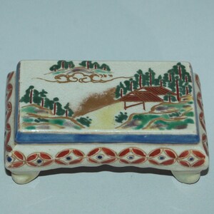 May include: A rectangular ceramic trinket box with a lid. The lid features a painted landscape scene with trees, a house, and a path. The box has a blue border and red and blue decorative accents.