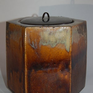 May include: Hexagonal ceramic container with a dark brown lid and a small handle. The container has a glazed finish with shades of brown, tan, and hints of green. The shape is geometric and the overall aesthetic is rustic.