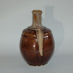 Puede incluir: Una jarra de cerámica marrón con cuerpo redondeado y cuello estrecho. La jarra tiene una superficie texturizada con líneas verticales y un pico de color claro. La jarra es probablemente hecha a mano y tiene un aspecto rústico.