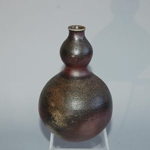 Peut inclure: Un vase en céramique fait à la main en forme de gourde avec une surface texturée et tachetée. Le vase présente une palette de couleurs marron foncé et rouge-violet, avec un col étroit et une base arrondie. Le vase est exposé sur un support transparent.