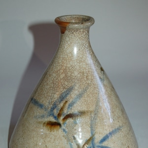 May include: A ceramic vase with a crackle glaze. The vase has a conical shape with a narrow neck and a brown rim. Blue and brown bamboo-like designs are painted on the off-white surface.