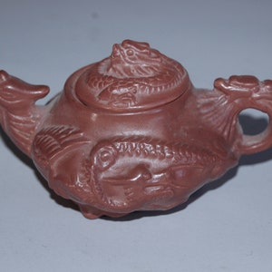 Puede incluir: Una tetera pequeña de arcilla roja con un diseño de dragón. La tetera tiene una tapa con un dragón en la parte superior y un asa de dragón.