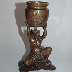 Op de afbeelding: Bronzen sculptuur van een knielende figuur die een klein, rond vat ondersteunt. Het vat heeft bloemsnijwerk. De figuur is gedetailleerd met gelaatstrekken en kleding. De sculptuur staat op een voet.