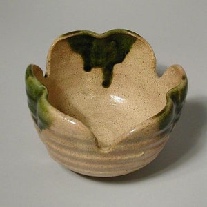 May include: A ceramic bowl with an unusual, organic form. The bowl has a light beige interior and exterior with a textured, ridged pattern. The rim and accents are a dark green colour. The bowl is likely handmade and suitable for decorative purposes.