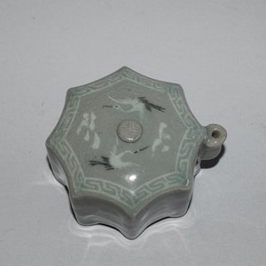 May include: A celadon-colored, star-shaped ceramic container with a lid. The lid features white crane and symbol designs, with a Greek key border. A small loop is on the side. The container is likely for storing small items.