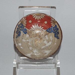 Puede incluir: Un pequeño cuenco de cerámica con una banda superior roja decorada con flores blancas similares a margaritas. La sección inferior muestra una base beige con detalles de volutas azules y patrones dorados en espiral, con un borde marrón oscuro.