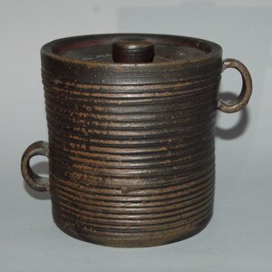 Puede incluir: Un recipiente cilíndrico de cerámica hecho a mano con tapa. La olla marrón oscuro presenta líneas horizontales texturizadas y dos asas de bucle. La tapa tiene un pequeño pomo cilíndrico. El borde interior de la olla es de un tono marrón más oscuro.