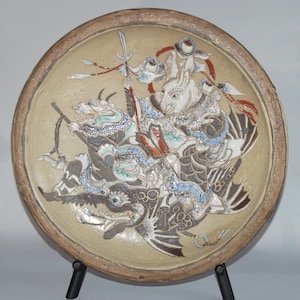 Könnte beinhalten: Beige Keramikplatte mit einem erhabenen Reliefdesign eines Kaninchenkriegers, der einen Drachen reitet. Der Krieger hält ein Schwert und ist von anderen Fabelwesen umgeben. Die Platte hat einen braunen Rand und steht auf einem schwarzen Ständer.