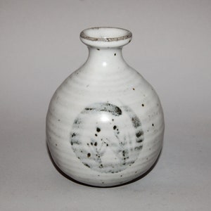 Puede incluir: Un jarrón de cerámica blanco con cuerpo redondo y cuello estrecho. El jarrón tiene una superficie texturizada con motas negras y un diseño circular. El borde es de color marrón claro.